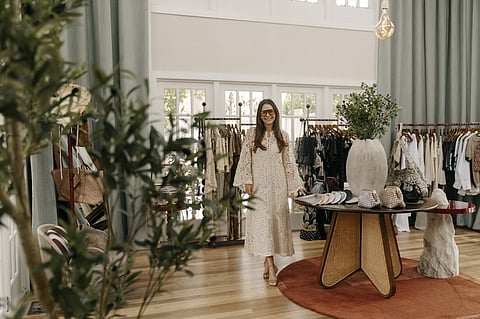 Adriana Castro in a cream lace dress beside a round table of handbags in a bright boutique