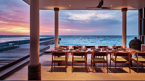 A long dining table set on a covered terrace facing the ocean at sunset