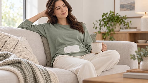 Woman relaxing on a couch with an abdominal heat patch and mug.