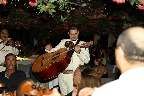 A guitarist in a white mariachi suit plays beside diners under flowering vines