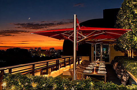 Rooftop terrace with red umbrellas and dining tables under an orange sunset sky