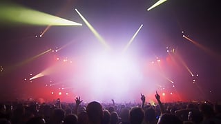 Silhouetted concertgoers raise their hands beneath yellow spotlights and stage lighting.