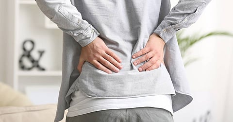 A young adult holding their lower back feeling chronic pain in a modern living room full of natural lighting.