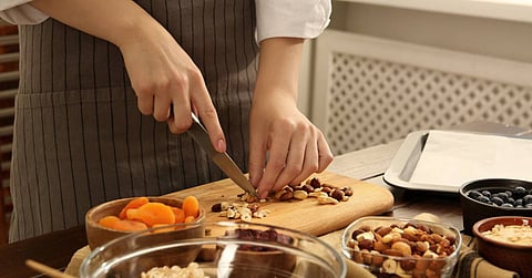 A person wearing a striped apron chops mixed nuts on a wooden cutting board, with bowls of dried apricots and other food nearby.