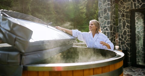 Woman wearing a white robe outdoors taking the plastic round cover from a hot tub while steam comes out.