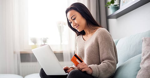 A young woman sitting cross-legged on her couch holds an orange credit card in one hand while using a laptop.