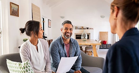 A man and woman sit on a couch and look at a woman sitting opposite. The woman holds a tablet displaying graphs.