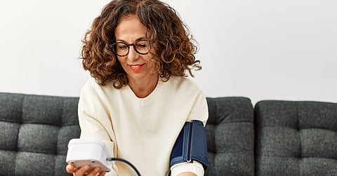 A woman sits on a grey couch and looks at a device in her hand. She has a blood pressure cuff around her left bicep.