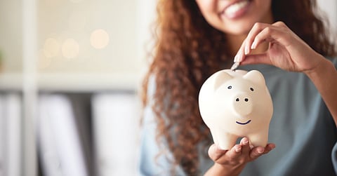 A woman smiles as she puts a coin into a white piggy bank, which is also smiling. She wears a gray shirt.