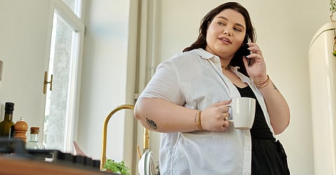 A woman with black hair standing in her kitchen and talking on a smartphone while holding a white coffee mug.