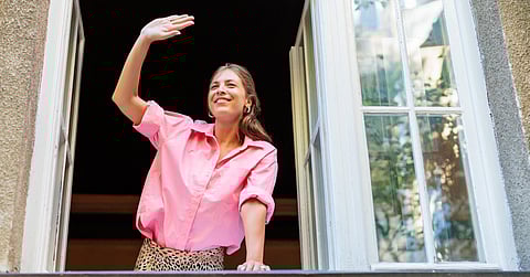A woman waves from the window of her home while smiling. She is wearing a pink shirt and leans on a railing.