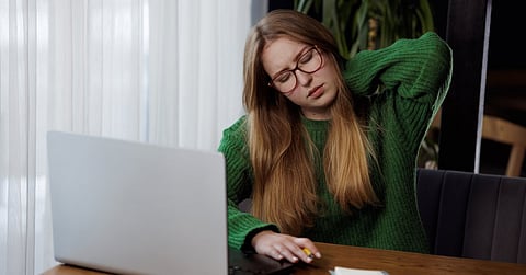 A woman sits in front of a laptop while holding her neck and closing her eyes. She tilts her head toward the left.