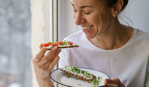 A close-up view shows a woman eating a piece of toast with a smear of cream cheese, tomatoes, and microgreens.