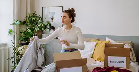 A woman picks up a pair of gray sweatpants from her bed, where other clothes and two donation boxes sit.