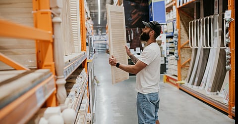 A man wearing a white shirt and jean shorts holds a wooden shutter. He is surrounded by different pieces of lumber.