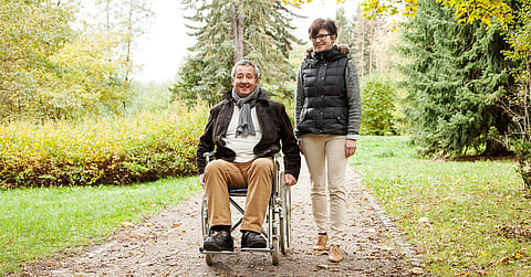 A woman walking alongside a man in a wheelchair. Both are dressed in warm clothes while on an outdoor path.