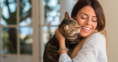 A woman wearing a light-blue blouse is cuddling a brown Tabby cat. She has her eyes closed and is smiling.
