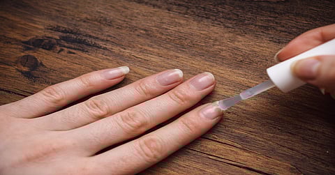 A woman paints her own nails. One hand rests on a brown wooden table, while the other applies nail polish.