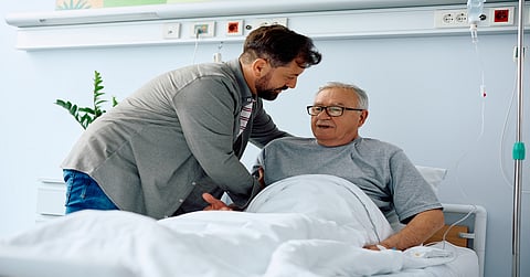 An elderly man sits upright in a hospital bed. A younger man puts his hand behind the elderly man's back for support.