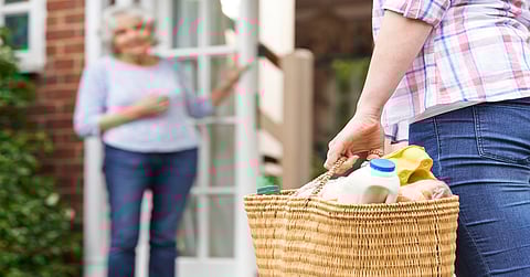 A woman carrying a wicker bag filled with groceries walking toward a home where an older woman waits by the door.