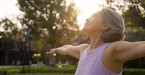 A woman with grey hair tilts her head up and grins while she performs a side arm raise stretch in a sunny outdoor setting.