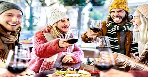 A group of friends wearing scarves and hats toasts with glasses of red wine over a charcuterie board.