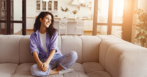  A woman relaxes on a beige couch in a bright living room with natural sunlight streaming through open glass doors.