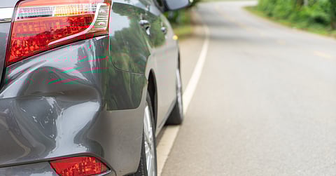 A close-up of the back bumper of a silver car with a dent on the right side, parked on the side of the road.