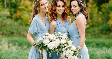 Three bridesmaids in light blue dresses stand outdoors, holding a white floral bouquet, with a blurred outdoor background.