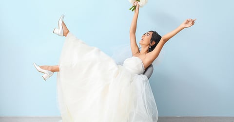 A woman in a strapless wedding dress sits and leans back in the air, lifting her feet and hands up while holding a bouquet.