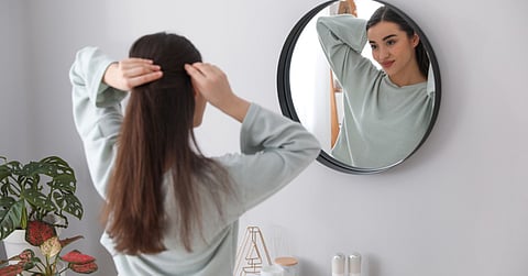 Young woman styling her hair while looking into a round mirror in a bright modern bedroom or bathroom interior.