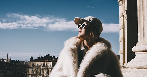 Stylish woman wearing sunglasses and a fur coat standing on a balcony overlooking a historic European city skyline.