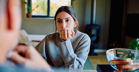 A woman sits at a kitchen table, listening intently to someone nearby, with food on a nearby counter.