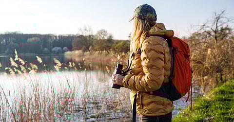 A woman stands on the side of a river, holding a pair of binoculars. She wears a winter coat and a backpack.
