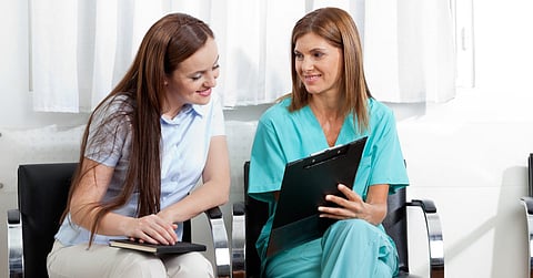 Two people are sitting in waiting room chairs. The dentist wearing scrubs holds up a clipboard while the woman reads it.