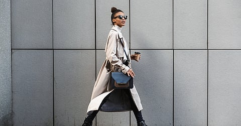 Stylish woman in beige coat strides along tiled gray wall, carrying coffee and purse, wearing sunglasses and boots.