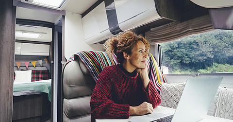 Woman using a laptop at a table inside a camper van, with a bed behind her and trees outside the window.