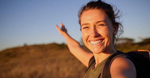 A woman traveling solo on a sunset hike. She smiles big at golden hour with her arm stretched out behind her.