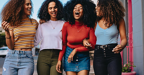 Four friends smile and laugh on a city sidewalk, wearing bright tops and jeans with arms around one another.