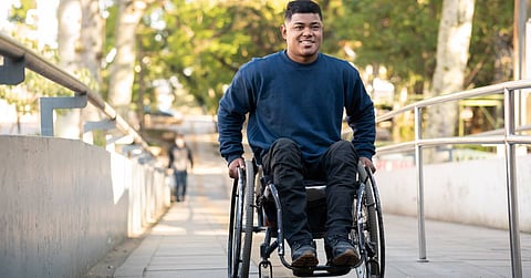 A man in a wheelchair pushing himself up an access ramp. There are trees and a park area behind him.