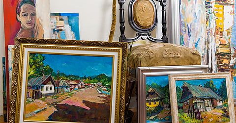 A collection of framed colorful paintings leaning on a floor beside tall canvases, a chair, and wall decor.