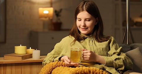 A woman smiles as she sits in her living room and enjoys tea. She has a blanket and sits next to lit candles.