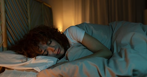 A woman with curly hair lies on her side in bed with the comforter pulled high up. Warm light glows in the background.