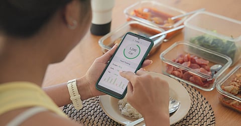 A young person sits at the dining table with food prep containers in front of them. They track calories on their cell phone.