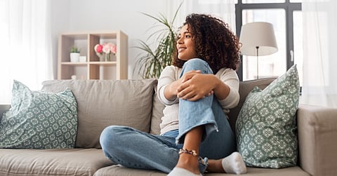 A young woman sitting on a couch with blue pillows in a well-lit home. She has her legs folded up on the couch.
