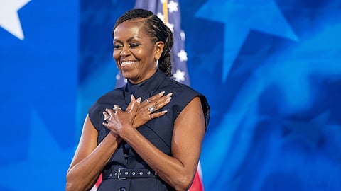 Michelle Obama stands smiling with her hands crossed over her chest in a gesture of gratitude. She wears a sleeveless navy blue dress with her hair styled in braids. Behind her, the American flag is partially visible, along with a blurred blue background featuring white stars.