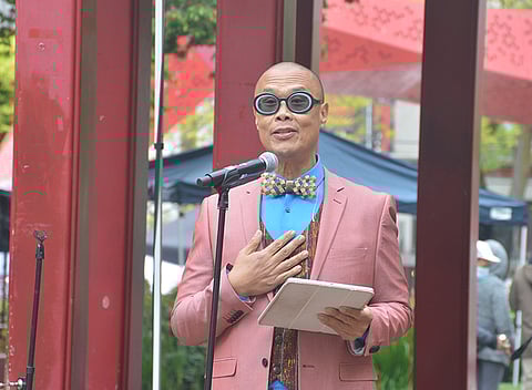 A man wearing a pink suit, blue shirt, and checkered bow tie speaks into a microphone at an outdoor event. He is holding a tablet in one hand and has the other hand placed over his chest. He also wears large, round sunglasses and stands in front of red pillars, with canopies and people in the background.