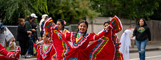 A young girl in a vibrant red traditional Mexican folkloric dress smiles as she dances in a parade. Her dress has colorful stripes and white lace trim, and she holds the skirt out wide as she twirls. Other dancers in similar attire are seen in the background, along with spectators watching from the sidewalk.