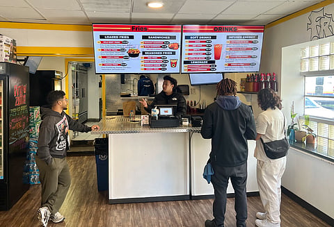 Three customers stand at the counter of FryZone, ordering food from the cashier. Above them, two digital menu screens display various fries, sandwiches, drinks, and energy options. The interior features wood flooring, a refrigerator stocked with beverages, and a chalkboard menu on the left.