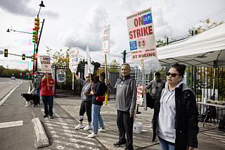 A group of workers stands on a sidewalk holding signs that read "On Strike at Boeing" during a labor protest. The signs also display "Aerospace Machinists" and "Unfair Labor Practices." Some of the protesters wear jackets and sunglasses, and one person holds a dog. A white tent with supplies is set up behind them, and the scene takes place near a road with a traffic light visible in the background under a cloudy sky.